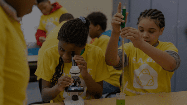 Two students in yellow tshirts, one looking through microscope, one using an injector